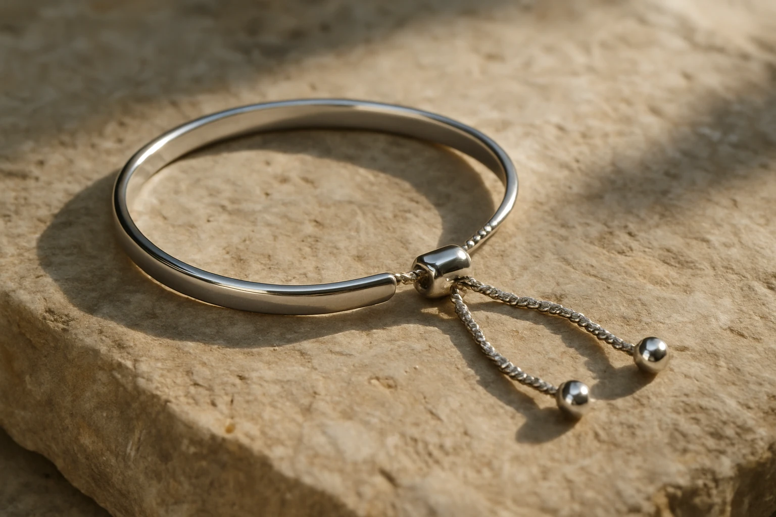 A close-up image of an elegant adjustable minimalist silver bracelet resting on a textured stone surface, with soft natural sunlight highlighting its sleek design and the delicate adjustment mechanism.